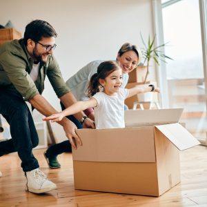Happy little girl with arms outstretched having fun being pushed by her parents in carton box at new home